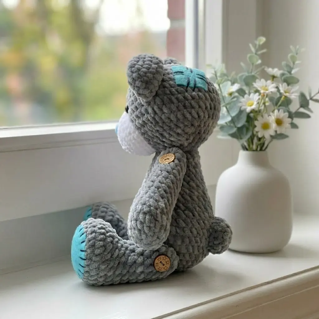 Side view of a gray chenille amigurumi bear with blue felt patches and wooden buttons sitting on a white windowsill.