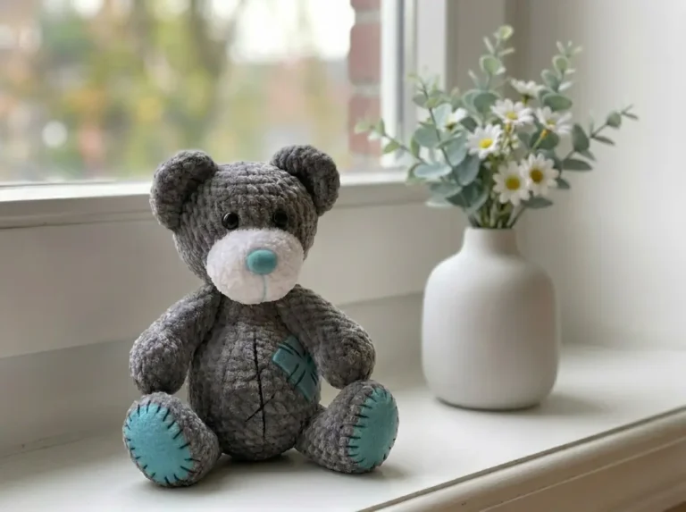 Grey plush amigurumi bear with a teal nose and patches sitting on a white windowsill next to a vase of daisies.