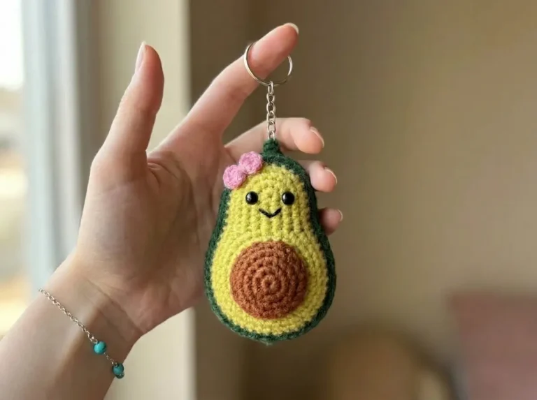 Hand holding a single crocheted avocado keychain amigurumi with a cute pink bow and safety eyes against a bright background.