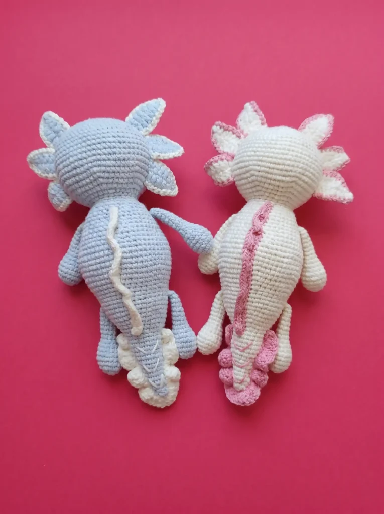 Two cute crocheted axolotl amigurumi toys in pastel colors displayed from the back on a pink background.
