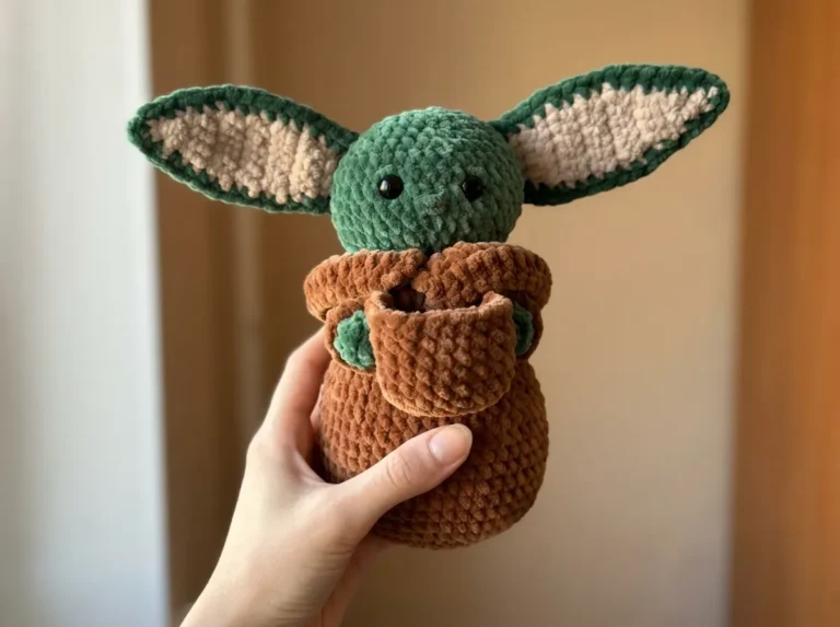 A hand holds a handmade velvet Baby Yoda amigurumi with green skin, brown robe, and beige ears against a plain indoor wall.