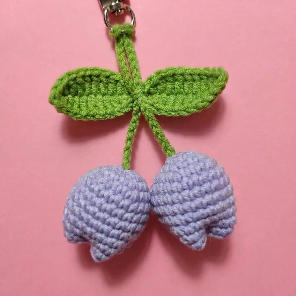 Close-up of a purple amigurumi keychain tulip with green leaves and a gold clasp on a pink background.