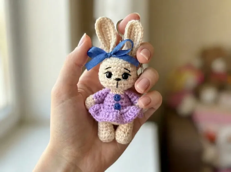Hand holding a beige crochet bunny keychain pattern in a purple dress and blue bow.
