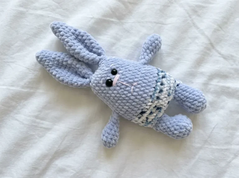 Light blue plush amigurumi bunny with variegated blue pants lying on a white wrinkled sheet.