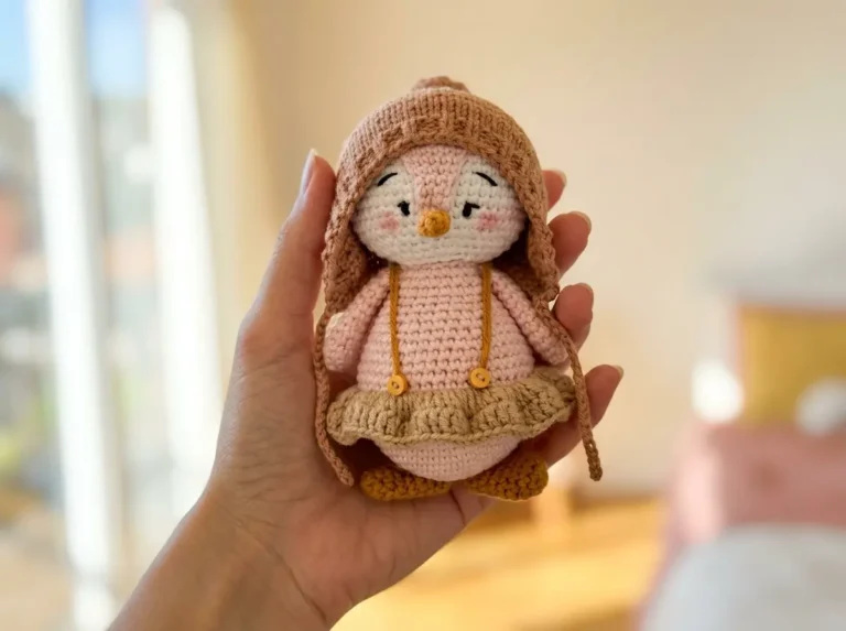 Hand holding a pink and white amigurumi penguin wearing a brown knit hat and ruffled skirt.