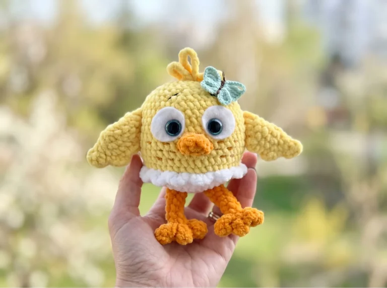 Hand holding a yellow plush yarn baby chick bird amigurumi with orange legs, white ruffle, and a blue bow in a bright room.