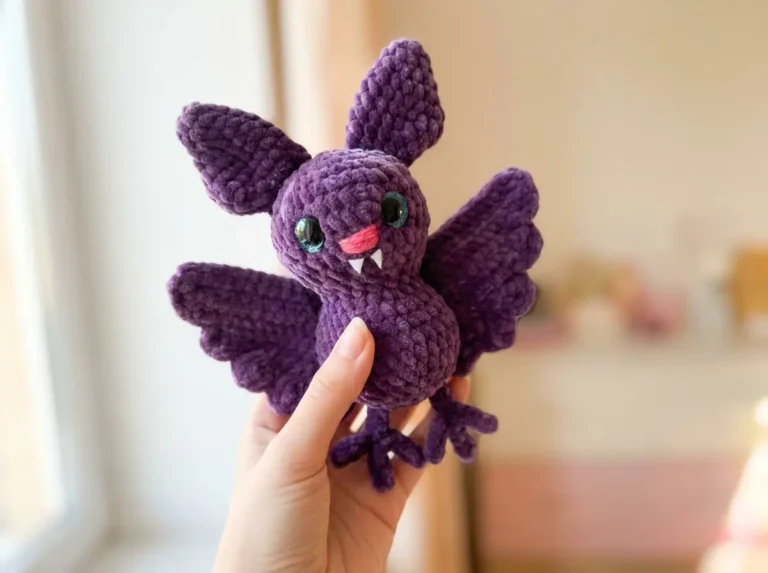 Hand holding a purple plush toy from a bat crochet pattern free, featuring a pink nose and fangs against a bright window.