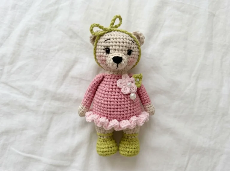 Crocheted bear amigurumi in a pink ruffled dress and green boots lying flat on a white fabric background.
