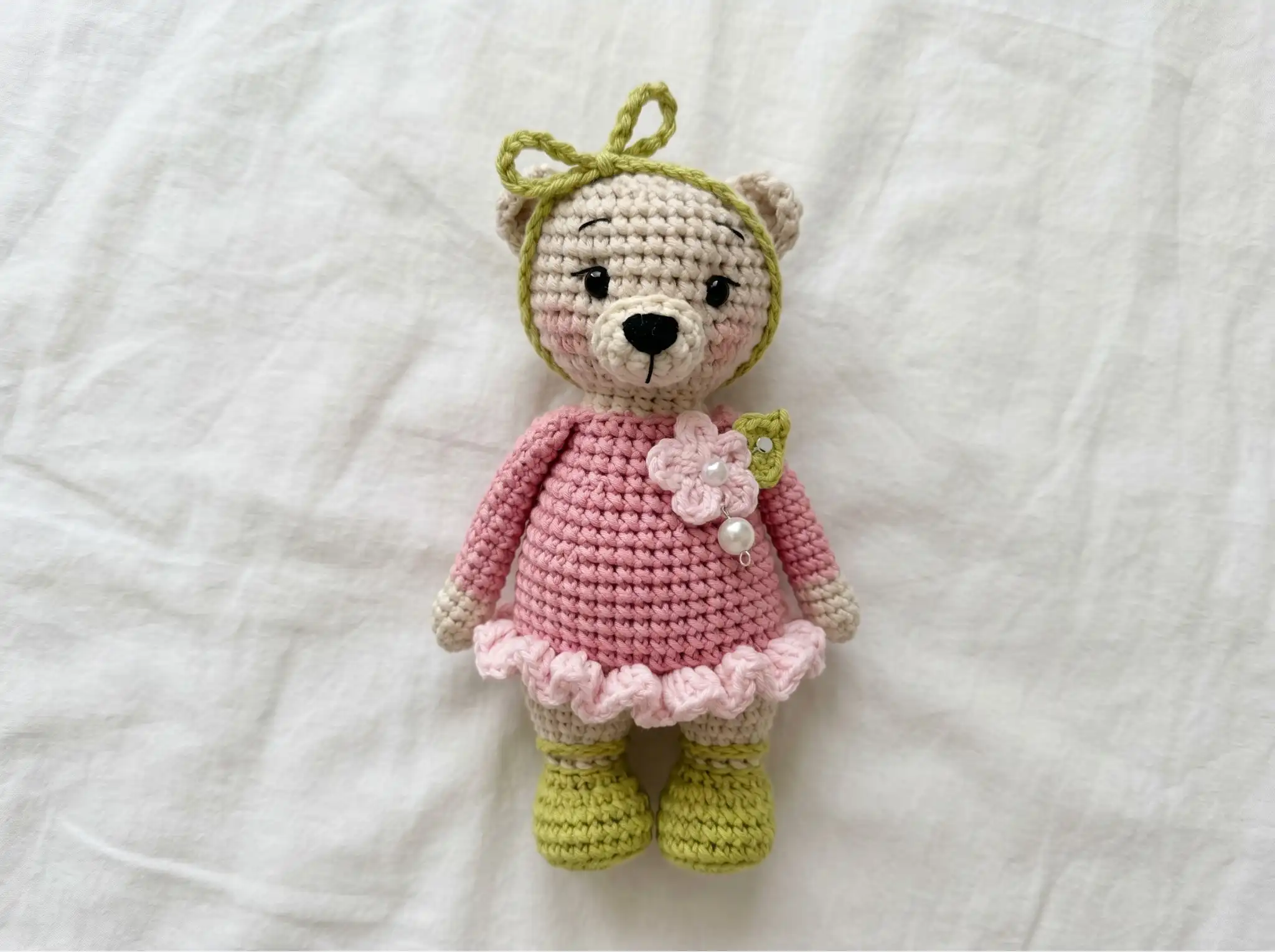 Crocheted bear amigurumi in a pink ruffled dress and green boots lying flat on a white fabric background.