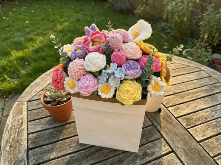 Colorful amigurumi bouquet with pink roses and white daisies in a paper bag on a sunny wooden table.