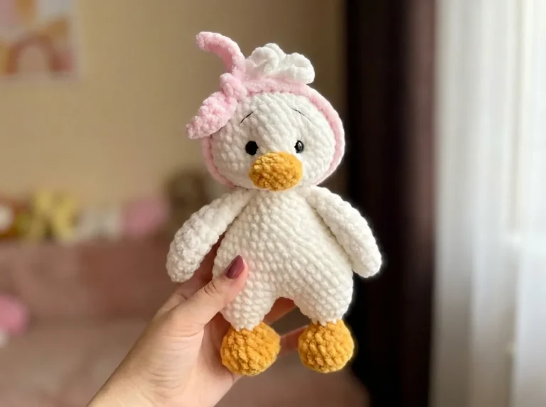 Hand holding a fluffy white toy with pink headband and orange beak from a crochet duck pattern free, with a room background.