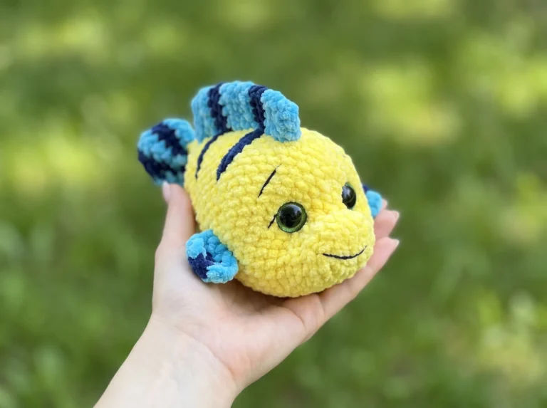 Hand holding a yellow and blue plush toy made using a crochet flounder free pattern in a sunny garden.