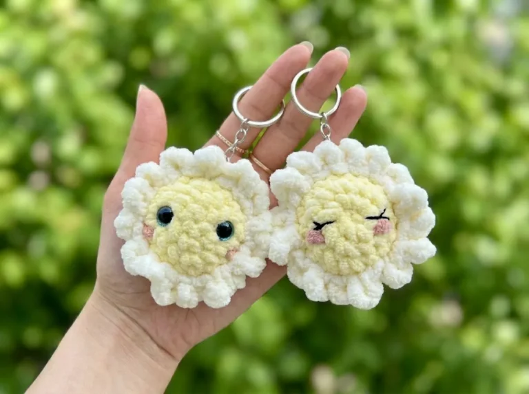 Hand holding two plush white and yellow crochet flower keychains made with velvet yarn against a green background.