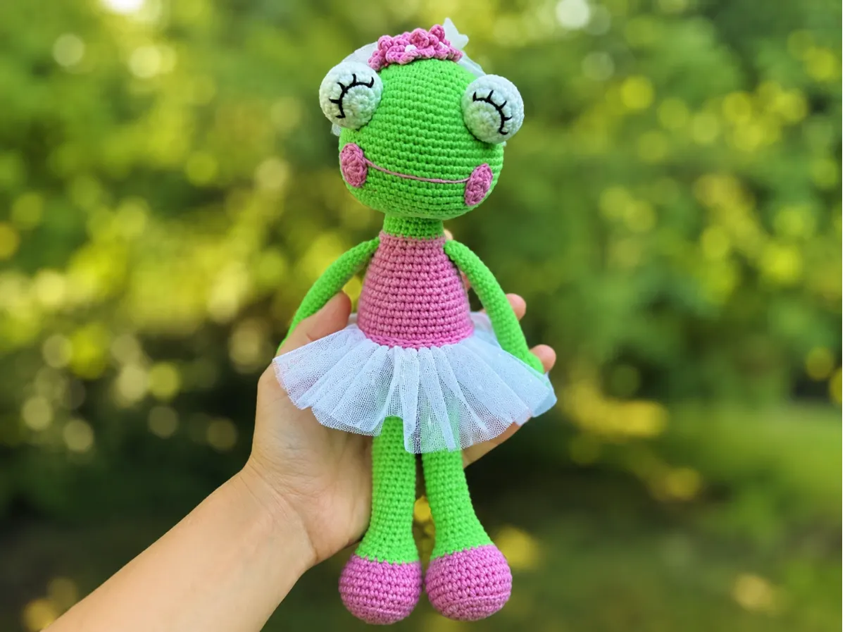 Hand holding a green crochet frog doll wearing a pink leotard and white tulle tutu against a blurry green background.