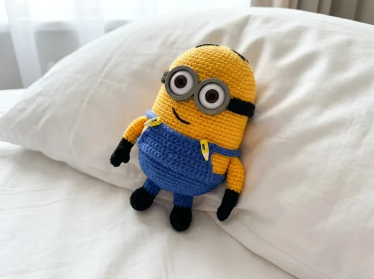 Yellow amigurumi minion in blue overalls and goggles on a white pillow. Crochet minion free pattern.