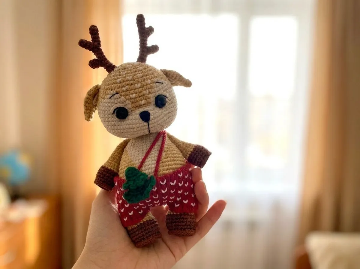 Horizontal view of a tan deer amigurumi with brown antlers and red patterned pants held against a bright window.