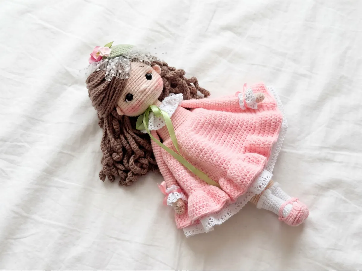 Doll amigurumi free pattern featuring a handmade toy with brown hair, pink dress, and lace hat lying on a white sheet.