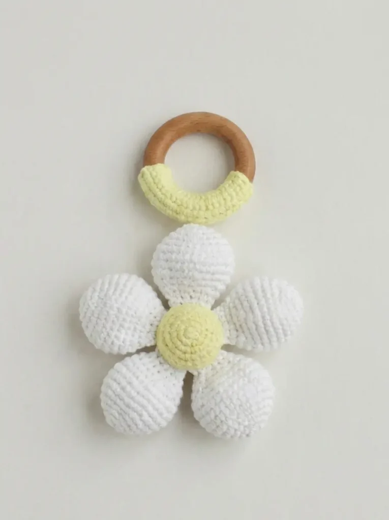 White and yellow crocheted flower rattle toy on a wooden ring, displayed against a plain neutral background.