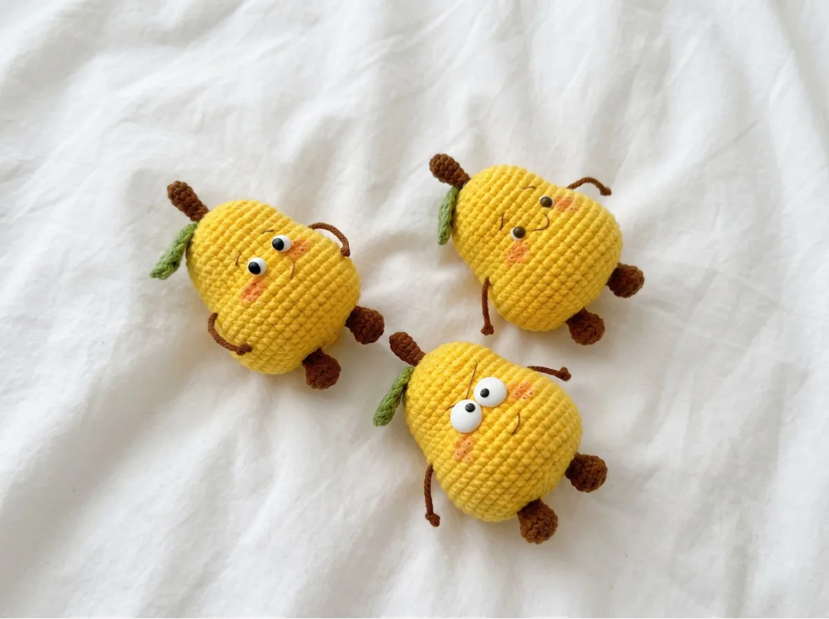 Three yellow amigurumi crochet pear figures with cute faces and brown stems on a bright white fabric background.