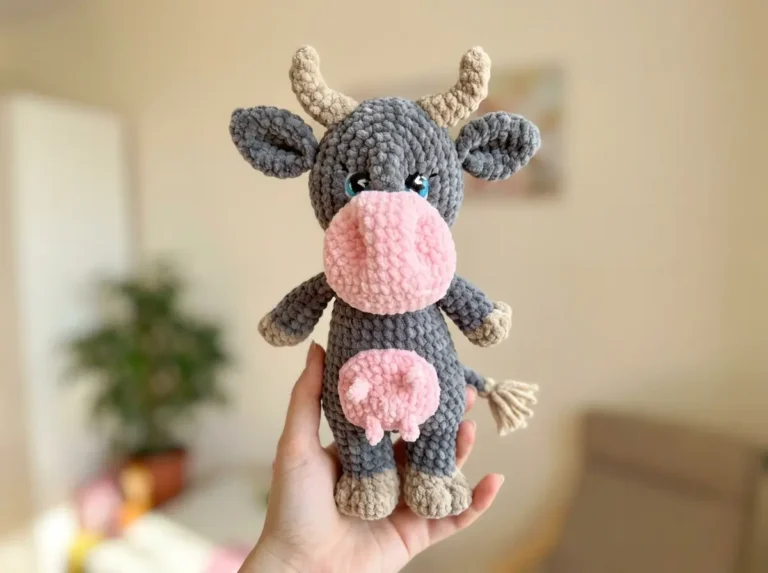 Hand holding a grey and pink plush velvet toy cow from a free cow crochet pattern amigurumi in a bright room.