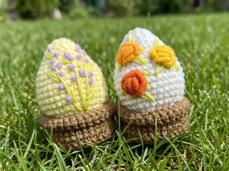 Two charming crochet easter egg creations in small brown pots sitting in a green lawn. One yellow with purple flowers, one white with orange.