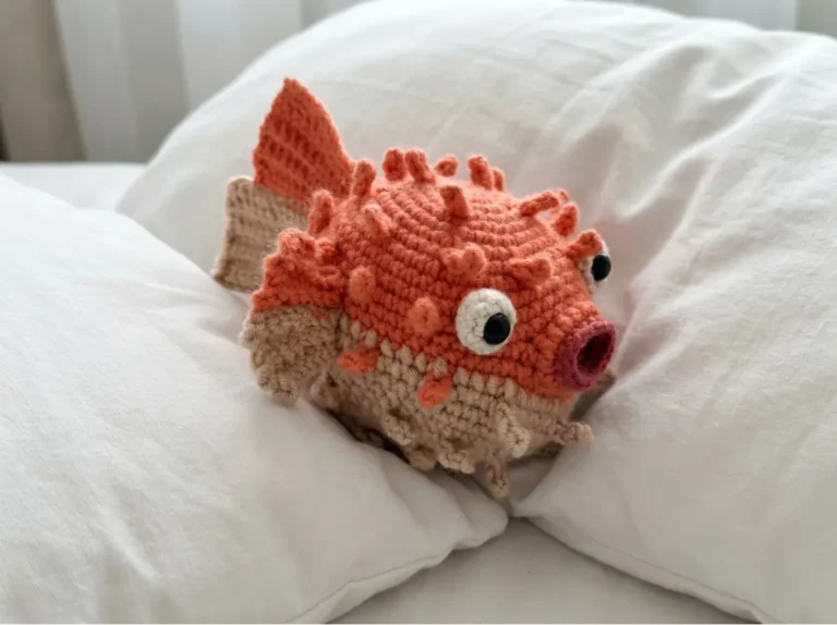 Orange and tan pufferfish crochet amigurumi toy with textured spikes, resting on soft white pillows.