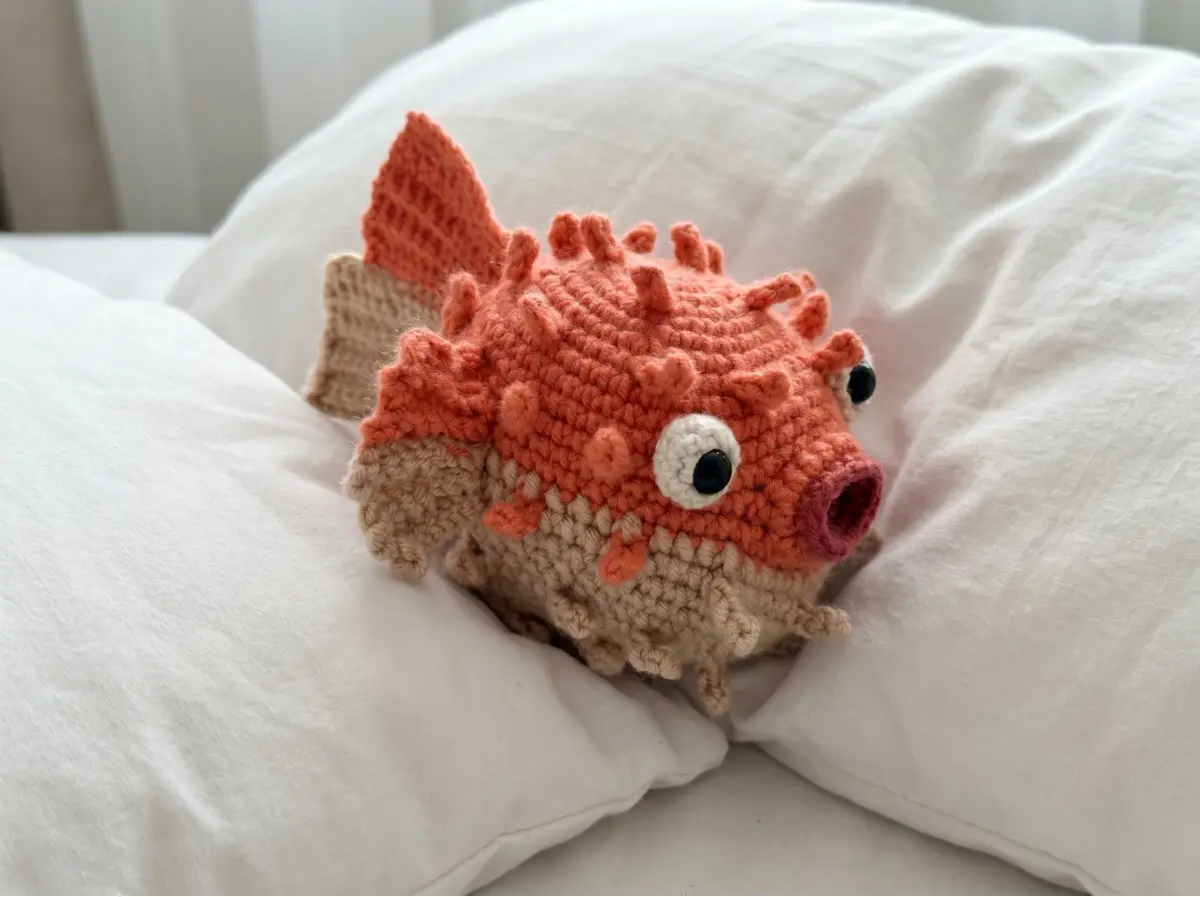 Orange and tan pufferfish crochet amigurumi toy with textured spikes, resting on soft white pillows.
