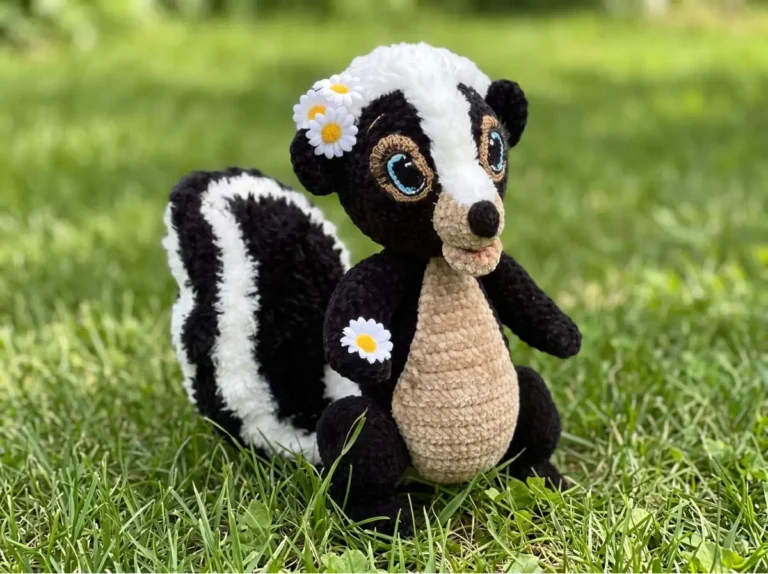 Black and white skunk crochet amigurumi with daisy flowers, sitting in green grass outdoors.