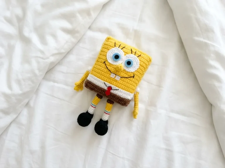 Yellow SpongeBob crochet toy with a red tie sitting on a wooden surface next to yellow yarn and a hook.