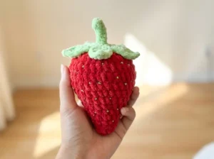 A hand holding a red plush strawberry amigurumi with green leaves and gold bead seeds against a blurred background.