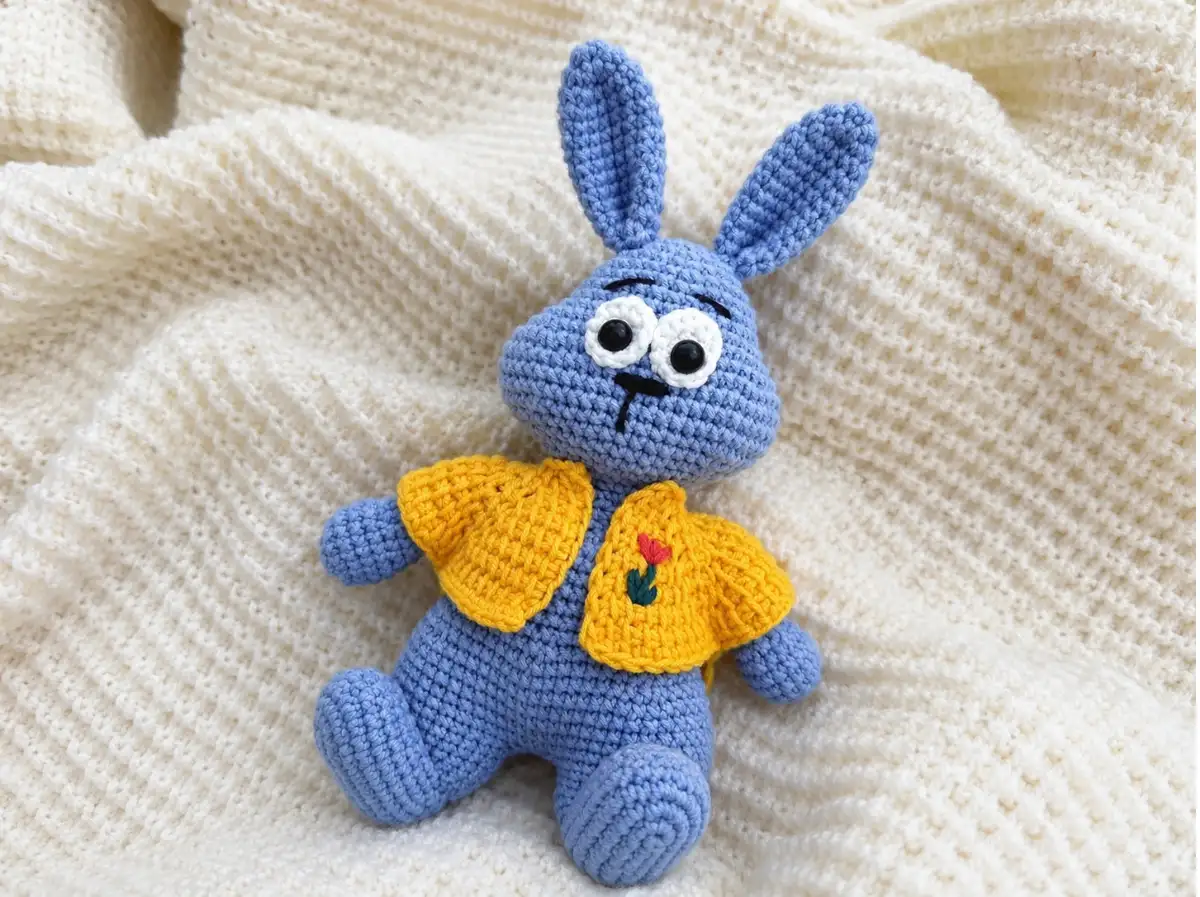 Light blue amigurumi bunny with floppy ears sitting on a cream textured blanket by a bright window.