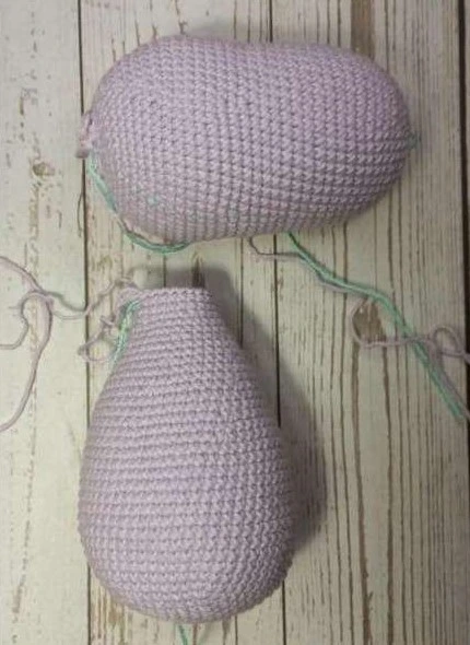 Finished separate pieces of a light lilac crocheted amigurumi unicorn pattern, including the stuffed oval head and teardrop-shaped body, laid out on light-colored wood.