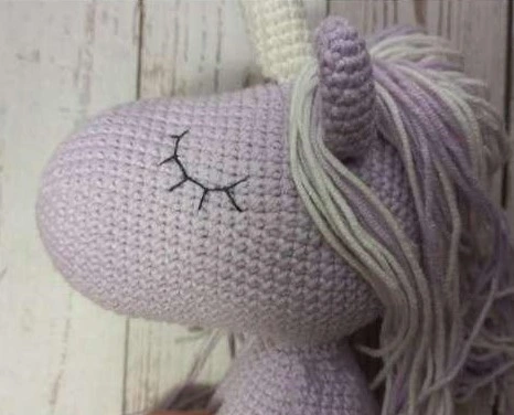 Profile view of a crocheted light lilac unicorn face featuring neat, black embroidered sleeping eyes with eyelashes, a lilac and cream yarn mane, and a textured horn.