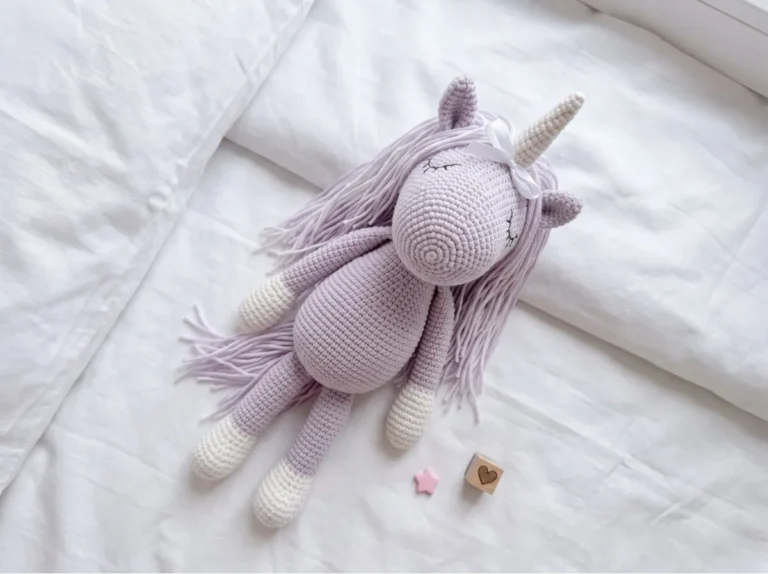 A light purple toy made from our free unicorn amigurumi pattern lying flat on white bedding with a wooden heart block.