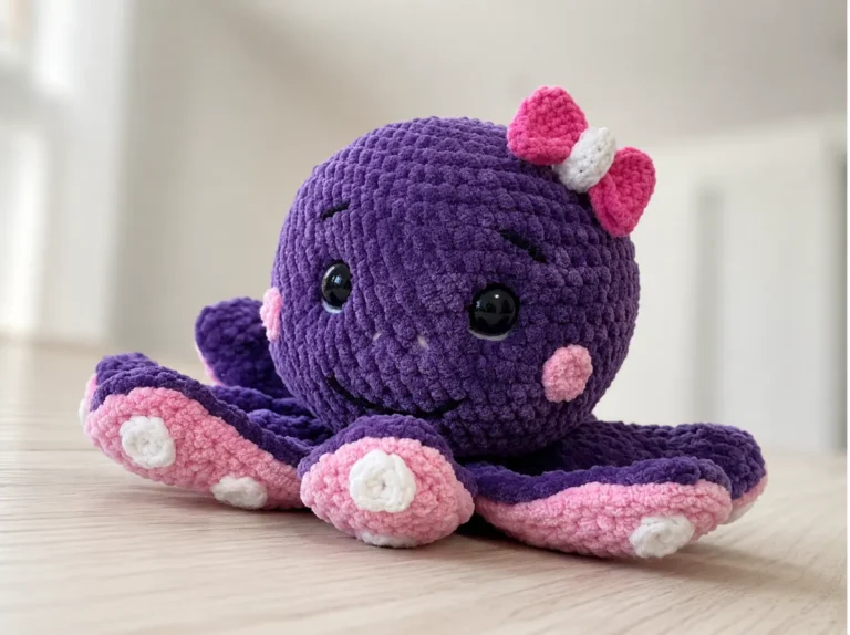Purple plush yarn octopus amigurumi pattern free sitting on a wood table, featuring pink tentacles and a pink bow