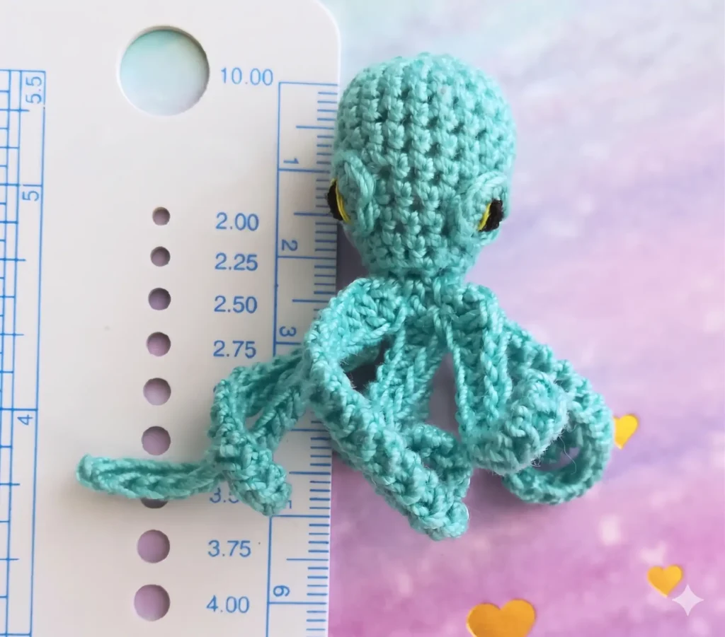 A small teal crocheted octopus placed next to a white measuring gauge on a pastel purple background with yellow hearts, showing its height at just under 2 inches.
