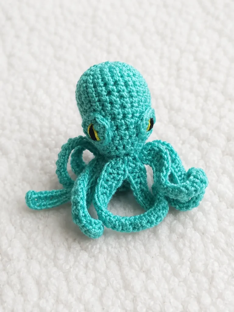A tiny teal crocheted octopus amigurumi sitting on a fluffy white surface, featuring eight curling tentacles and embroidered yellow and black eyes.