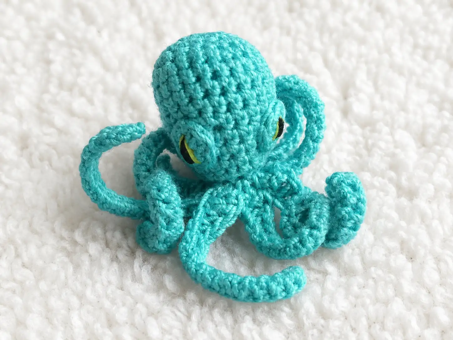 A slightly angled top-down view of a small teal crocheted octopus resting on a white textured background, highlighting its naturally curling tentacles and focused embroidered eyes.