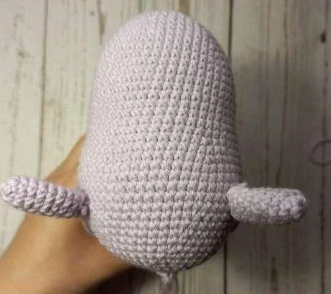 A hand holding a light lilac crocheted amigurumi unicorn body with two matching crocheted arms sewn on either side just below the open neck hole.