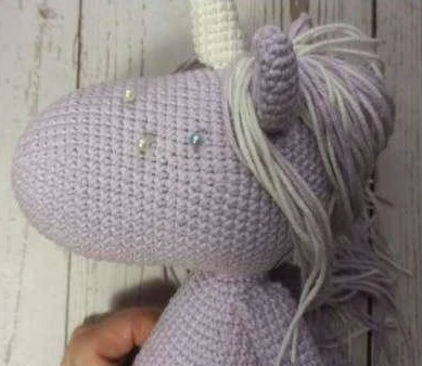 Crocheted light lilac unicorn head with colored straight pins placed on the face to map out symmetrical eye embroidery lines.