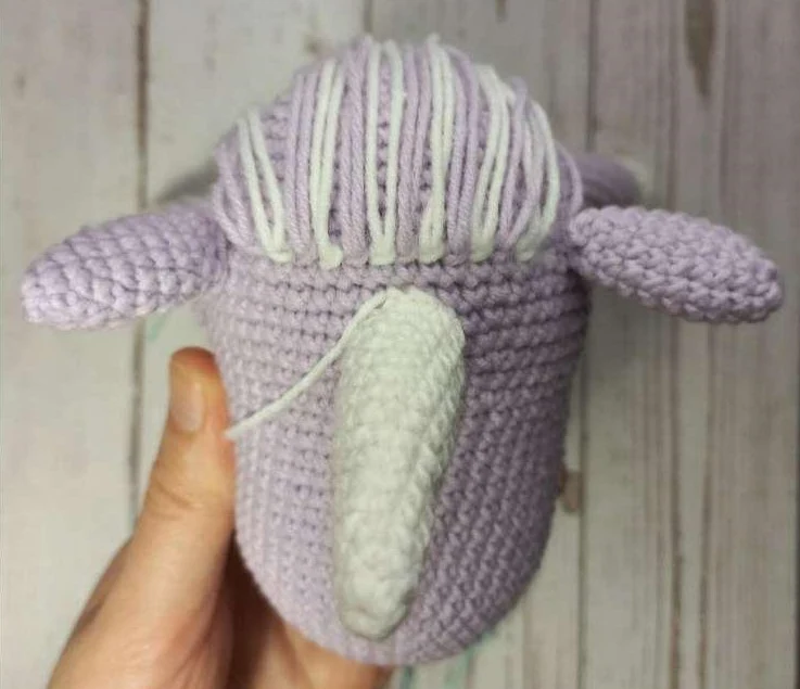Top-down view of a light lilac crocheted unicorn head showing a cream horn, two side ears, and alternating cream and lilac yarn strands neatly tied between the ears to form the mane.