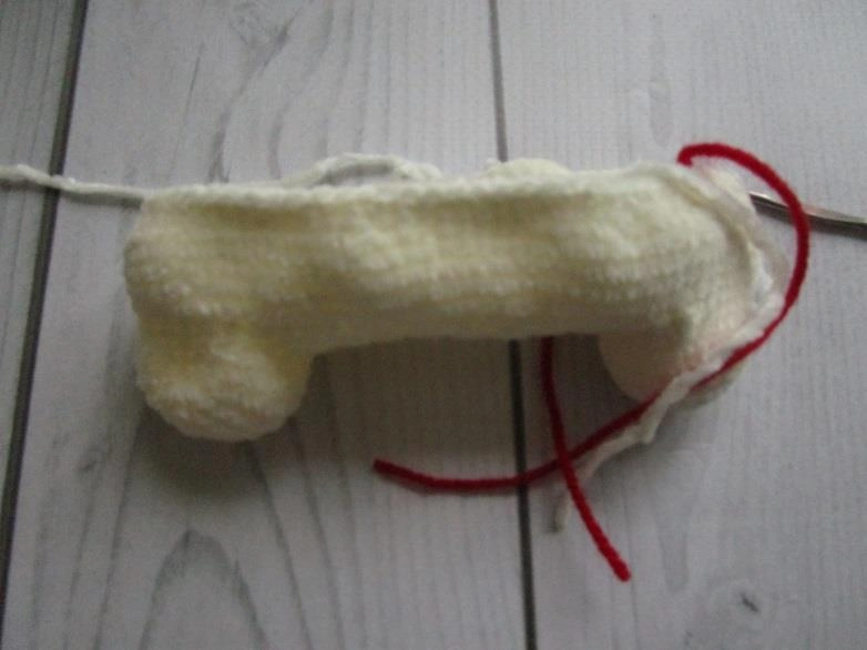 Two white bulky plush amigurumi legs connected by a flat crocheted bridge, featuring a piece of red yarn used as a stitch marker.