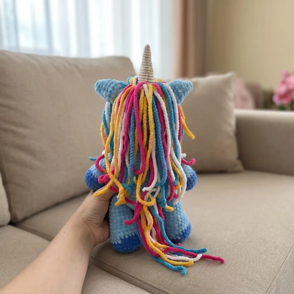 Back of blue plush crochet unicorn with long colorful yarn mane held on beige sofa. Amigurumi unicorn free pattern.