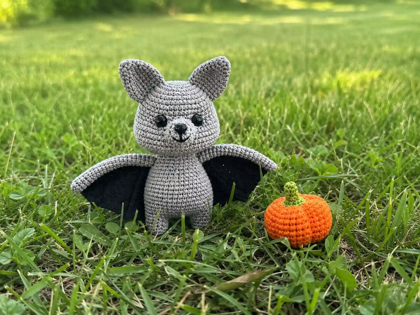 A small grey crochet bat with black wings sits on green grass next to a tiny orange pumpkin.