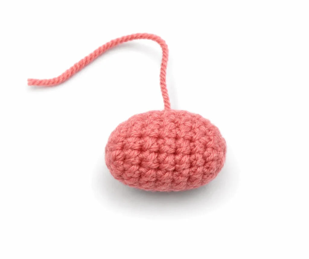 A completed oval crochet nose worked in coral yarn for the bucktooth bunny amigurumi, resting flat on a white surface with a long yarn tail.