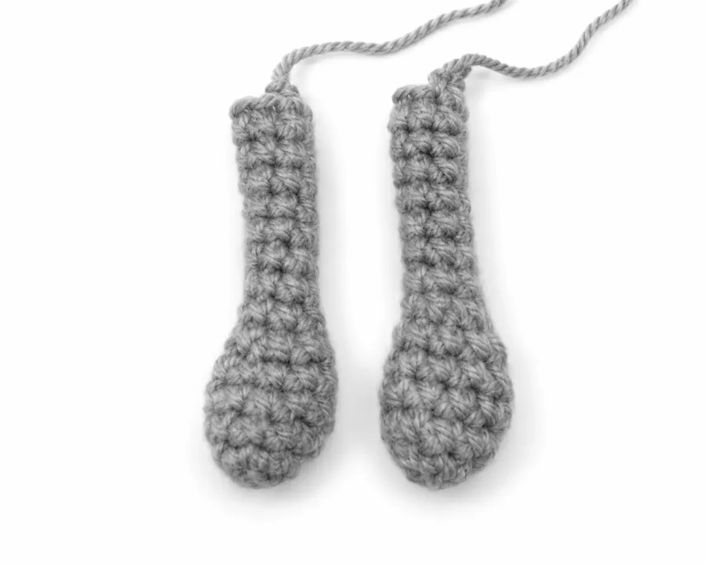 A pair of completed steel-gray crocheted arms for the bucktooth bunny amigurumi, laid flat against a white background with long yarn tails left for sewing.