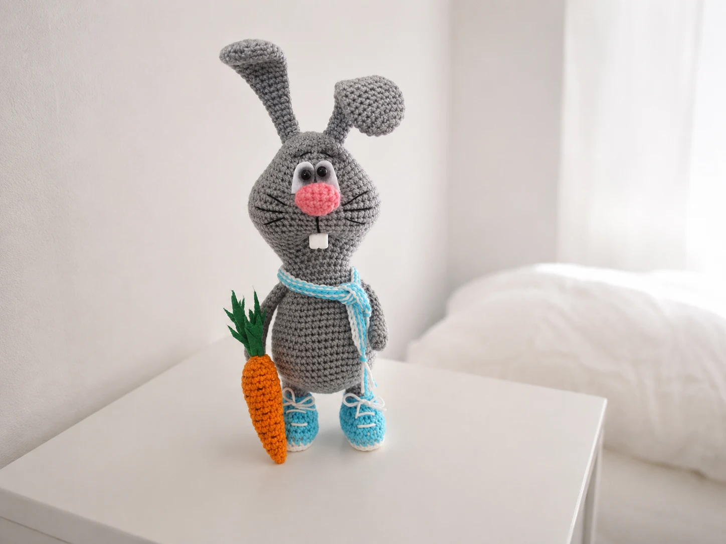 Grey crocheted bucktooth bunny amigurumi free pattern toy with a carrot, blue shoes, and scarf on a white table.