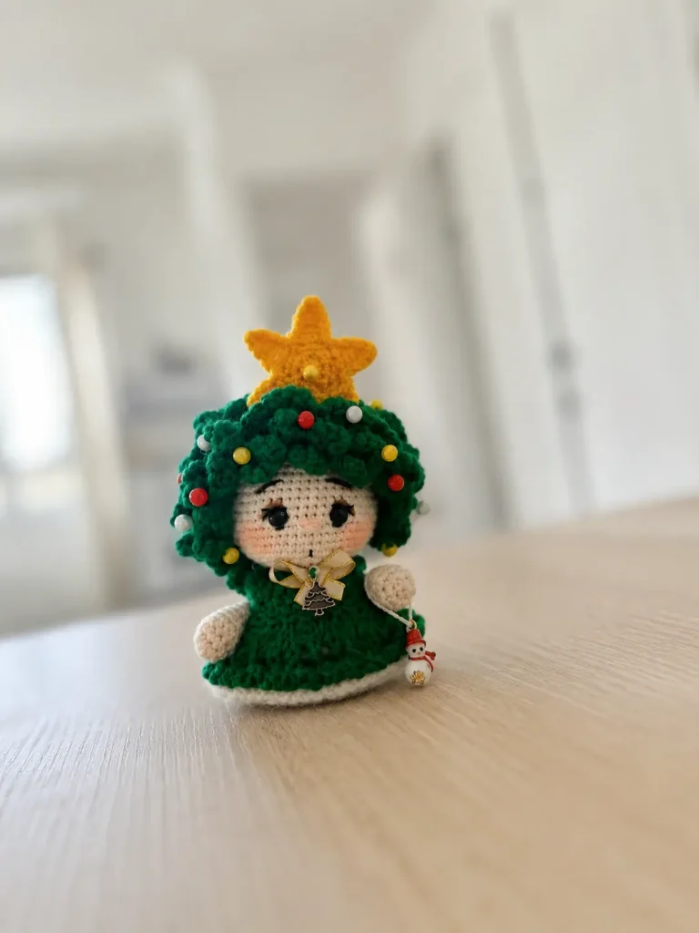 A finished, self-standing crochet Christmas tree amigurumi doll displayed on a light wood surface, showcasing the layered green wig with multi-colored beads, the gold star topper, the detailed dress, the button-jointed arm holding the tiny snowman accessory, and the complete embroidered facial expression.