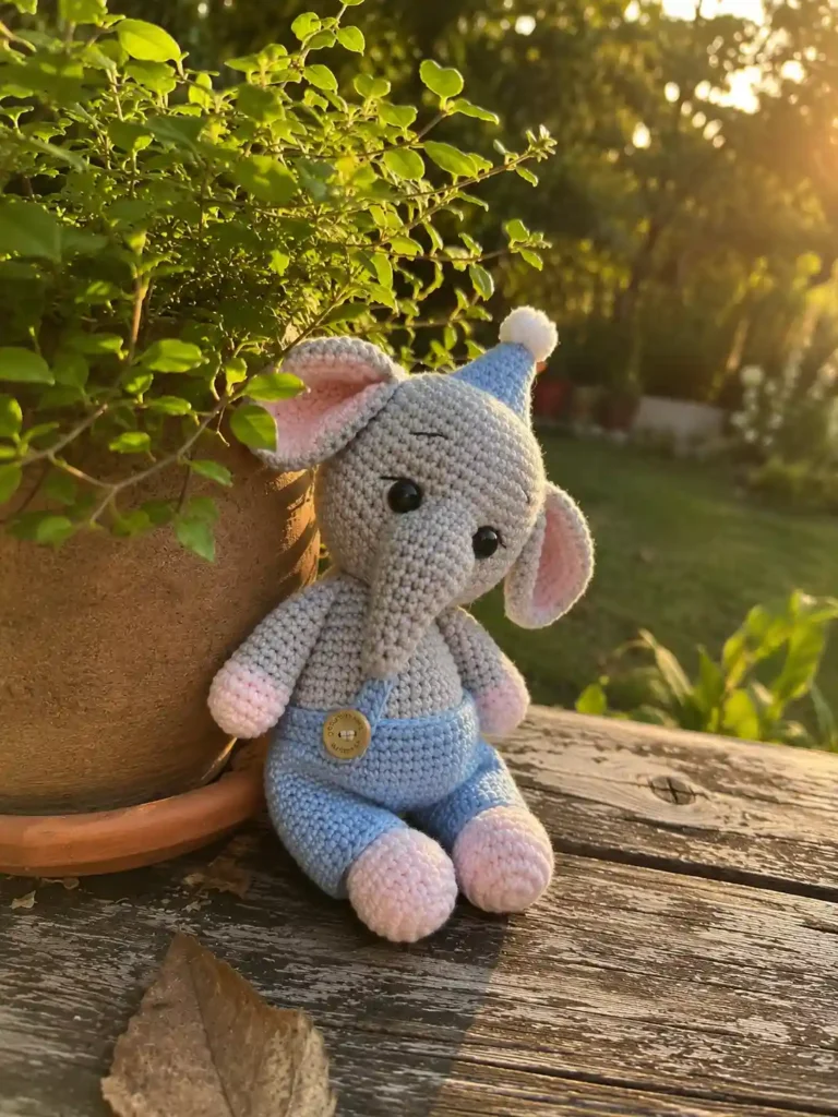 Crochet baby elephant amigurumi in dove gray with blush pink paws, blue pants with a button, and a matching party hat topped with a white pom-pom, sitting outdoors next to a potted plant.