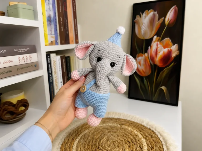 Hand holding grey crochet baby elephant amigurumi in blue overalls with books and tulip art in background